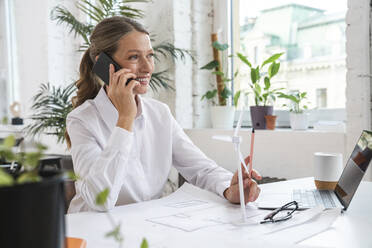 Mature businesswoman talking through smart phone with blueprint on table - VPIF07818