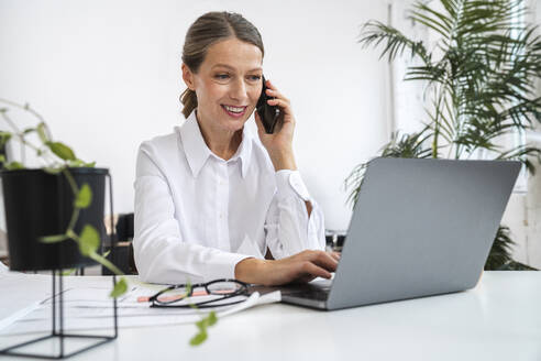 Lächelnde Geschäftsfrau, die über ein Mobiltelefon spricht und einen Laptop im Büro benutzt - VPIF07819
