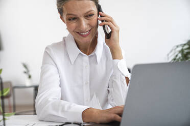 Glückliche reife Geschäftsfrau, die über ein Mobiltelefon spricht und einen Laptop im Büro benutzt - VPIF07820