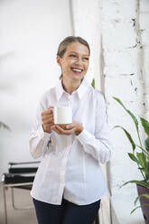 Happy mature businesswoman with coffee cup standing by wall - VPIF07842