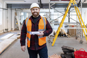 Happy engineer standing with rolled document at construction site - WPEF06952