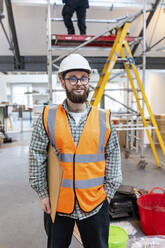 Lächelnder junger Ingenieur mit Klemmbrett auf einer Baustelle - WPEF06954