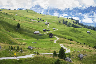 Hiker on footpath among green fields and wooden huts, Sass de Putia, Passo delle Erbe, Dolomites, South Tyrol, Italy, Europe - RHPLF23527