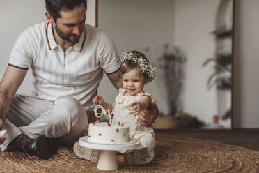 Father sitting with daughter touching birthday cake at home - MDOF00520