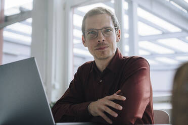 Smiling businessman with laptop sitting in office - JOSEF15810