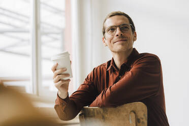 Lächelnder Geschäftsmann mit Einweg-Kaffeebecher auf einem Stuhl im Büro sitzend - JOSEF15822