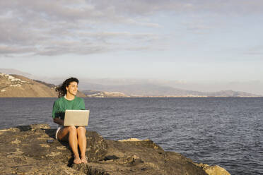 Lächelnde Frau mit Laptop auf einem Felsen am Meer sitzend - LJF02463