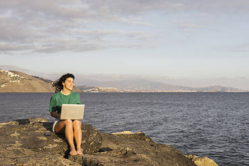 Smiling woman with laptop sitting on rock by sea - LJF02463