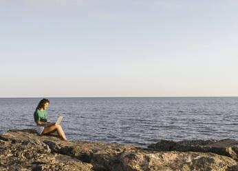 Frau arbeitet am Laptop auf einem Felsen am Meer sitzend - LJF02464