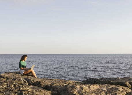 Woman working on laptop sitting on rock by sea - LJF02464