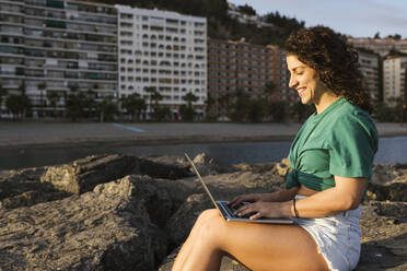 Lächelnder Freiberufler mit Laptop auf einem Felsen am Strand sitzend - LJF02465