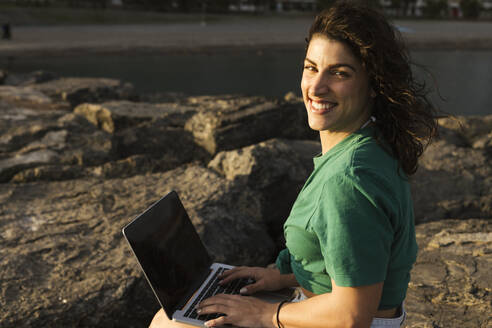 Happy woman with laptop sitting on rock at beach - LJF02466