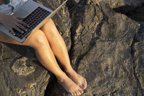 Woman using laptop sitting on rock at sunny day - LJF02467