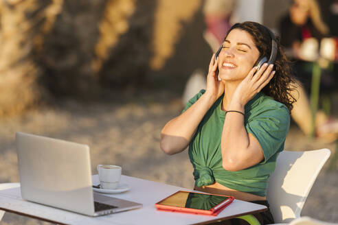 Happy woman wearing wireless headphones listening to music at outdoor cafe - LJF02470