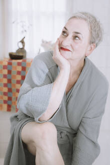 Thoughtful senior woman with hand on chin sitting in bathroom - NGF00768