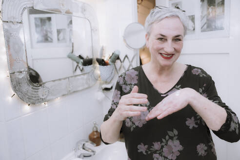 Happy woman applying moisturizer cream on hand in bathroom - NGF00770