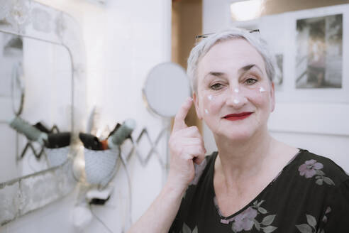 Smiling woman applying cream on face in bathroom - NGF00771