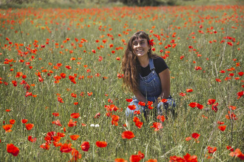 Glückliche junge Frau sitzt inmitten eines Feldes mit Mohnblumen - PCLF00221