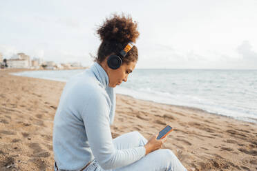 Frau, die ein Smartphone benutzt und über Kopfhörer am Strand Musik hört - JOSEF15856