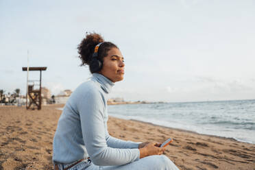 Nachdenkliche junge Frau sitzt mit Smartphone am Strand - JOSEF15857