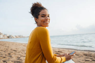 Glückliche Frau mit Mobiltelefon am Strand sitzend - JOSEF15862