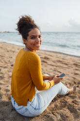 Glückliche junge Frau sitzt mit Handy am Strand - JOSEF15863