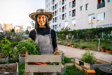 Smiling woman holding crate of carrots in urban garden - GDBF00014