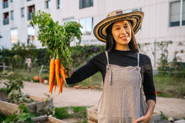 Glückliche Frau mit Karotten in einem städtischen Garten stehend - GDBF00017