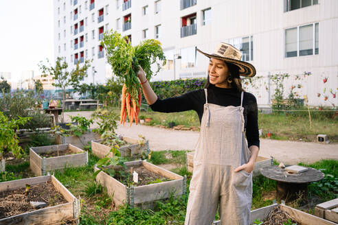 Happy woman standing with carrots in urban garden - GDBF00018