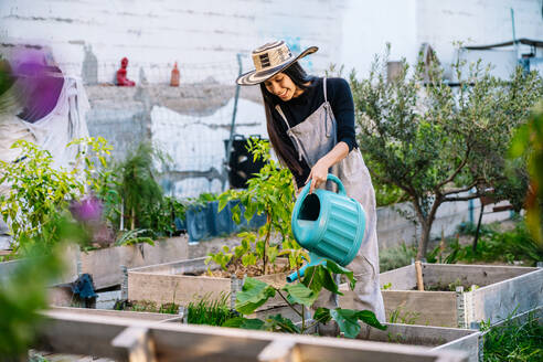 Lächelnde Frau mit Hut bewässert Pflanzen im städtischen Garten - GDBF00021