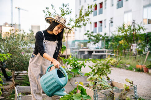 Happy woman wearing hat watering plants with can in urban garden - GDBF00023