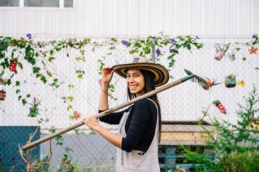 Happy woman wearing hat holding rake in urban garden - GDBF00031