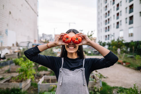 Happy woman holding tomatoes over eyes in urban garden - GDBF00034