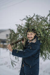 Lächelnder Mann trägt Tannenbaum im Schnee - ANAF00902