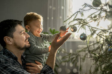 Happy father and son decorating Christmas tree at home - ANAF00909
