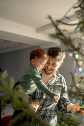 Smiling son and father decorating Christmas tree at home - ANAF00910