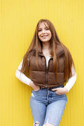 Glückliche junge Frau mit langen Haaren lehnt an einer gelben Wand - DAMF01183