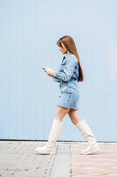 Junge Frau benutzt ein Smartphone und geht an einer blauen Wand vorbei - DAMF01187