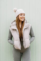 Happy woman wearing knit hat standing in front of green wall - DAMF01194