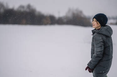 Smiling boy wearing knit hat standing in snow - ANAF00920