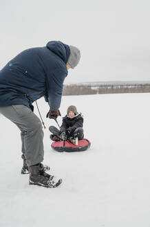 Vater zieht seinen Sohn auf einem aufblasbaren Schlitten im Schnee sitzend - ANAF00937