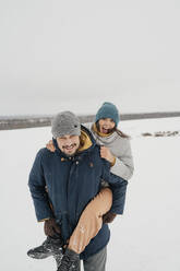 Happy man giving piggyback ride to woman in snow - ANAF00939