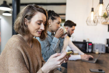 Smiling businesswoman using mobile phone with colleagues in background - WPEF06968