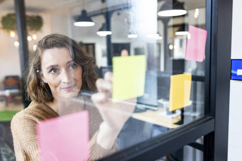 Smiling businesswoman sticking adhesive notes seen through glass - WPEF06997