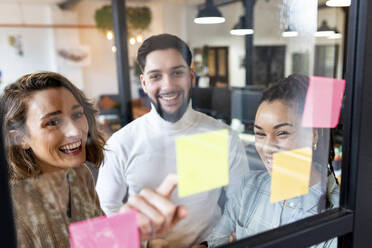 Smiling businesswoman with colleagues reading adhesive notes on glass in office - WPEF07002