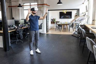 Junger Geschäftsmann mit Virtual-Reality-Simulator steht mit erhobenen Händen im Büro - WPEF07045