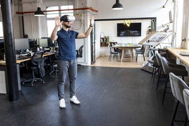 Young businessman wearing virtual reality simulator standing with hands raised in office
 - WPEF07045