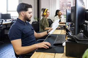 Young businessman using desktop and holding charts in office - WPEF07056