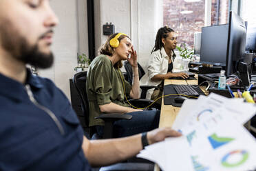Frustrierte Geschäftsfrau mit Kopfhörern vor einem Schreibtisch im Büro sitzend - WPEF07059