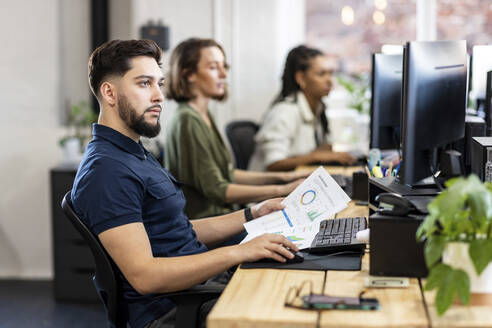 Junger Geschäftsmann mit Diagrammen, der an einem Desktop-PC im Büro arbeitet - WPEF07069